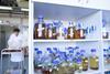 An image showing chemicals in bottles in a cabinet; a technician can be seen in the background pushing a trolley of glassware