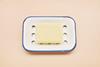 A photograph of a bar of yellow soap in a blue and white enamel soap dish, against a pink-orange background