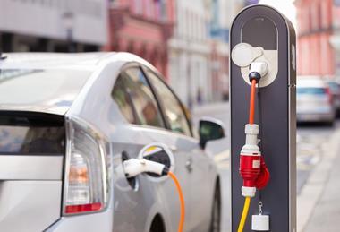 A photograph of an electric car connecting to a charging station at the side of a street